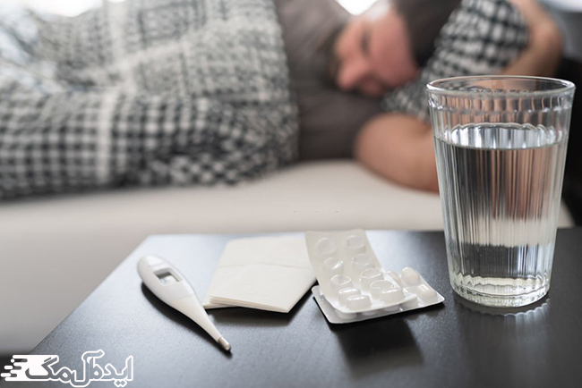مرد بیمار روی تخت خوابیده و یک لیوان آب، تعدادی دارو و تب‌سنج روبروی او قرار دارد