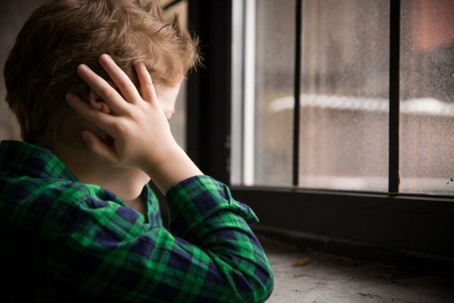 یک پسربچه مبتلا به اوتیسم کنار پنجره ایستاده و سر خود را با دو دست گرفته است