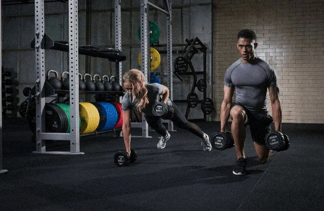 یک مرد و یک زن در باشگاه مشغول تمرین با دمبل و وزنه هستند
