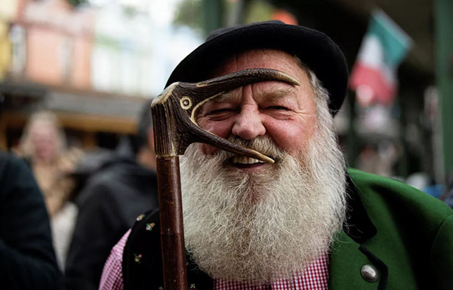 یک پیرمرد با ریش بلند و عصایی به شکل سر کلاغ رو به دوربین می خندد