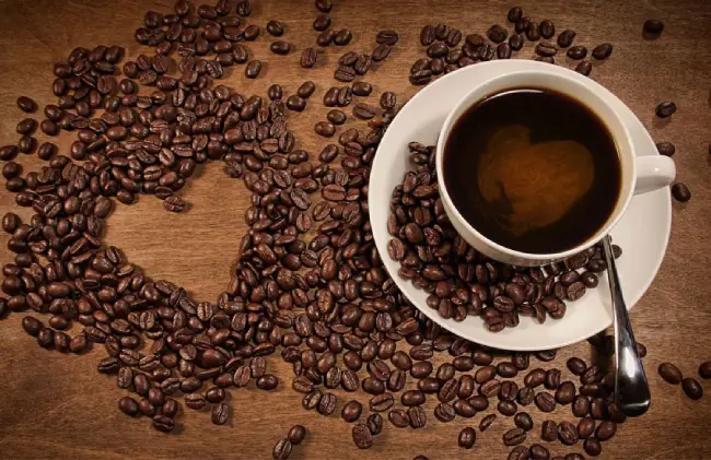 تعداد زیادی دانه قهوه روی میز ریخته شده و یک فنجان قهوه نیز روی میز قرار دارد