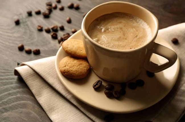 یک فنجان قهوه روی میز قرار دارد. داخل نعلبکی آن، دو کوکی گذاشته شده است و مقداری دانه قهوه روی میز ریخته شده است