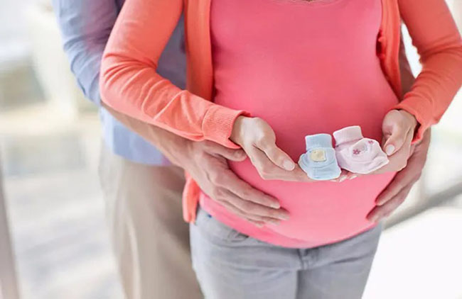 یک زن باردار و همسرش. زن دو جوراب یا کفش نوزاد به رنگ های صورتی و آبی را در دست نگه داشته و مرد، دستهایش را روی شکم زن گذاشته است