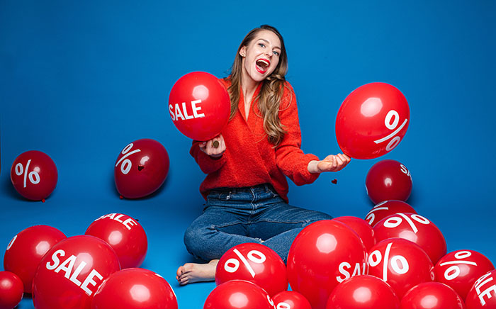 یک زن با پیراهن قرمز نشسته و بادکنک های قرمزی که روی آن نوشته شده «حراج» را به دست گرفته است و چند بادکنک قرمز هم دور و برش ریخته است